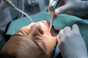 Man examining oral cavity of young African-American man working in in dental clinic with assistant.