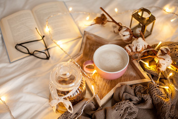 Mug of hot cappuccino on a wooden tray is on the bed. Cozy decor. Breakfast. Mug, plaid, cotton, candle. Book. Christmas lights. Holidays. Christmas. Autumn. Winter.