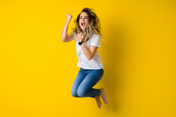 Fototapeta na wymiar Beautiful young blonde woman jumping happy and celebrating with raised hands and open mouth over isolated yellow background