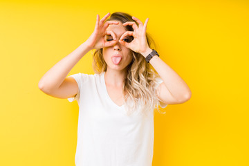 Young beautiful blonde woman over yellow background doing ok gesture like binoculars sticking tongue out, eyes looking through fingers. Crazy expression.