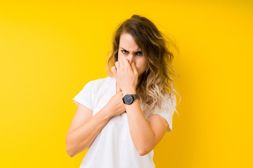 Young beautiful blonde woman over yellow background smelling something stinky and disgusting,...