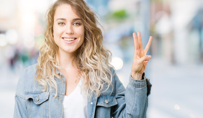 Beautiful young blonde woman wearing denim jacket over isolated background showing and pointing up with fingers number three while smiling confident and happy.