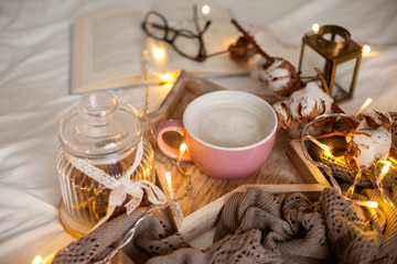Mug of hot cappuccino on a wooden tray is on the bed. Cozy decor. Breakfast. Mug, plaid, cotton, candle. Book. Christmas lights. Holidays. Christmas. Autumn. Winter.