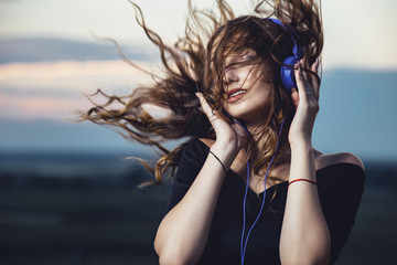 portrait of a beautiful girl in headphones with flying hair from the wind, young woman listening to music on the nature in the field