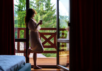 Woman in the morning holding a cup of tea or coffee and looking at the mountains standing near the window .