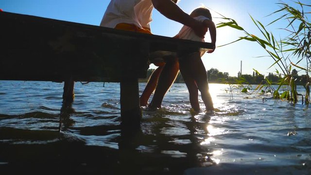 Father holding in hands his son while sitting on the edge of wooden jetty at lake and dangling his legs in water. Little child and his dad swinging feet in river at sunny day. Slow motion Close up