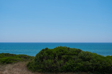Blue sky and ocean water behind green hill.