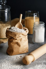 Bag of flour for preparing dough with sugar and other ingredients and kitchen utensils in a glass jars  in background. Food preparation on dark background.