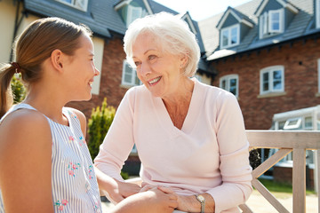 Granddaughter Sitting On Bench With Grandmother During Visit To Retirement Home