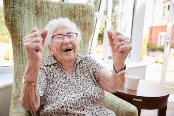 Portrait Of Excited Senior Woman Sitting In Chair In Lounge Of Retirement Home