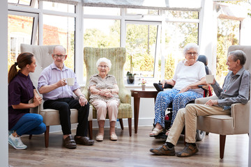 Male And Female Residents Sitting In Chairs And Talking With Carer In Lounge Of Retirement Home