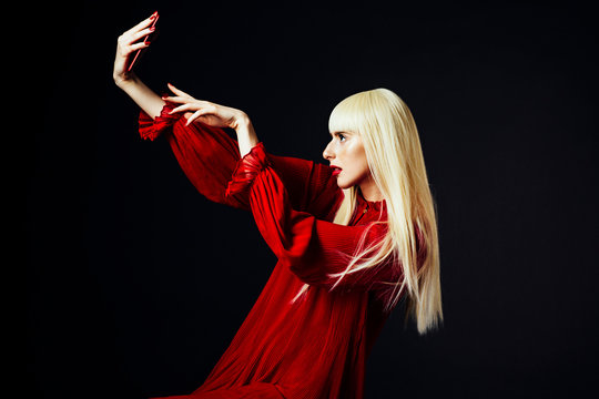 Portrait of a beautiful blonde woman holding a red smartphone about to press the button, isolated on black studio background