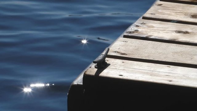Slow motion view of blue lake water with sunny highlights. Detail of wooden dock and metal boat cleat.