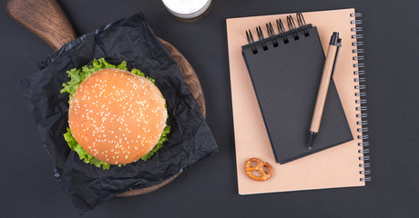 Big hamburger, beer and notebook for planning on a black background. Fast food and snacks. Free space for text. Flat lay. Copy space.