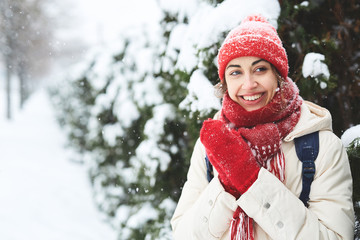 portrait of Cheerful smiling woman in white down jacket and red cap, scarf and mittens on the snowy alley with small spruces after blizzard in city. copyspace