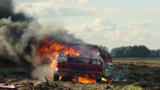The car burns in the field and its wheel explodes. Car on fire. Side View.