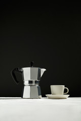 Coffee maker and white cup and saucer are on the white table. Black background blurred. Minimalism