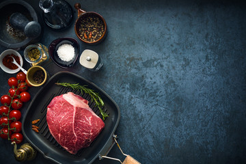 Raw marbled beef steak on a grill pan with salt, herbs and tomatoes. Top view and copyspace.