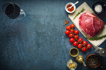 Raw marbled beef steak on dark concrete background with salt and herbs, tomatoes and glass of red wine. Top view and copyspace.