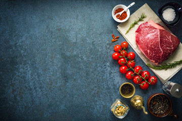 Raw marbled beef steak on dark concrete background with salt, herbs and tomatoes. Top view and copyspace.