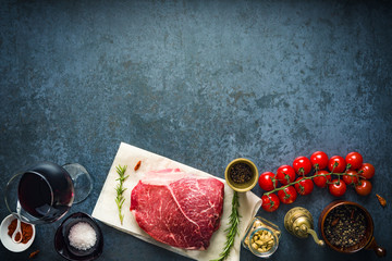 Raw marbled beef steak on dark concrete background with salt and herbs, tomatoes and glass of red wine. Top view and copyspace