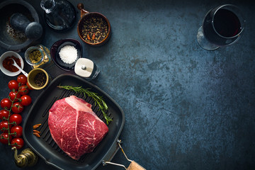 Raw marbled beef steak on a grill pan with salt, herbs and tomatoes and glass of red wine. Top view and copyspace.