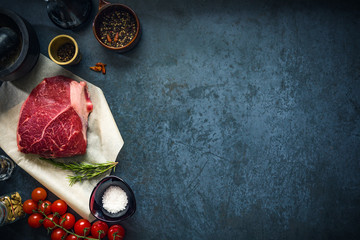 Raw marbled beef steak on dark concrete background with salt and herbs and tomatoes. Top view and copyspace.