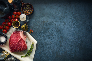 Raw marbled beef steak on dark concrete background with salt and herbs and tomatoes. Top view and copyspace.