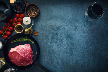Raw marbled beef steak on cast iron pan with salt and herbs, tomatoes and glass of red wine. Top view and copyspace.