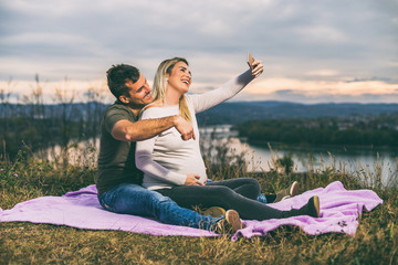 Happy husband and his pregnant wife taking selfie while they enjoy spending time together outdoor.