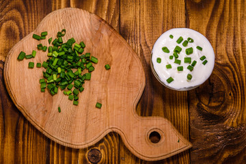 Glass bowl with sour cream and cutting board with chopped green onion on wooden table. Top view