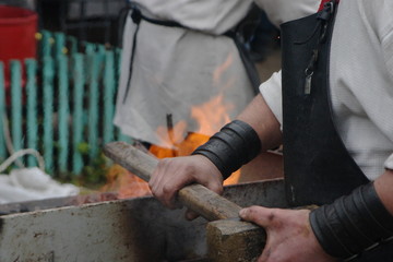 A blacksmith with a face not seen hold a hummer over a fire at an open blacksmith festival