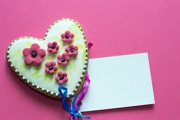 Gingerbread heart with white icing and pink flowers shit of paper on pink background. Love concept. Gift for womans and valentines day. Copy space for text.