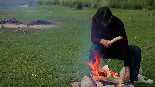 family vacation destination on the banks of the river in the mountains around the campfire. A middle-aged Caucasian man in a bard sweater playing with a white dog breed poodle. evening light 