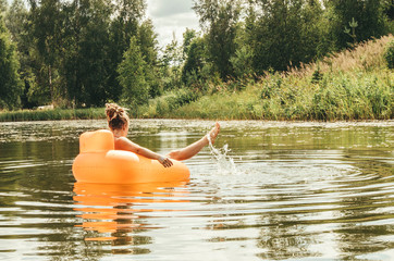 Young adult woman having fun, resting and sunbathing, relaxing on inflatable round orange chair toy and floating on lake pond in nature outdoors in summer.