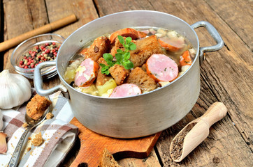 Garlic soup in an old saucepan with sausage, fresh garlic bulbs, cloves, colored pepper, wooden spoon, croutons and a towel on a rustic wooden table