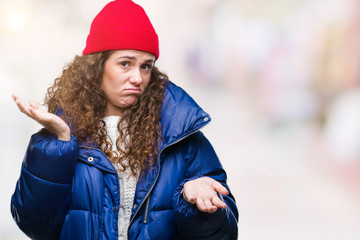 Beautiful young brunette curly hair girl wearing winter coat, wool cap and sweater over isolated background clueless and confused expression with arms and hands raised. Doubt concept.