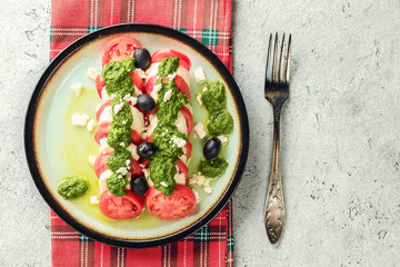 Tomato and mozzarella caprese salad and pesto sauce with olives on gray table served with red checked napkin, decanter with olive oil fork. Food italian appetizer