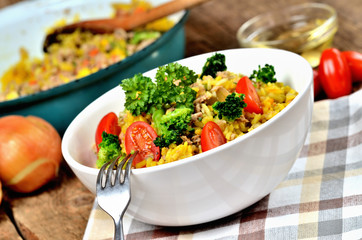 Close-up of fork and tuna risotto with vegetables, tomatoes, broccoli and parsley in the bowl, onions, oil and pan in the background