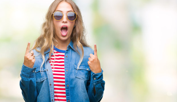 Beautiful young blonde woman wearing sunglasses over isolated background amazed and surprised looking up and pointing with fingers and raised arms.