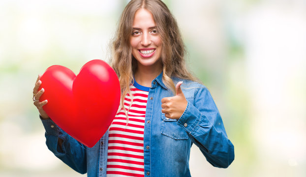 Beautiful young blonde woman holding heart valentine over isolated background happy with big smile doing ok sign, thumb up with fingers, excellent sign