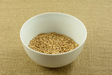Uncooked raw farro wheat in white bowl on textured burlap