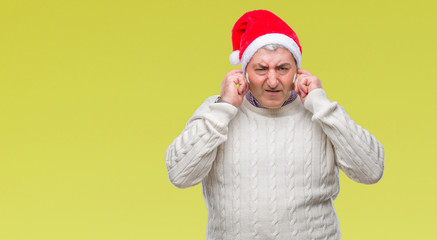 Handsome senior man wearing christmas hat over isolated background covering ears with fingers with annoyed expression for the noise of loud music. Deaf concept.