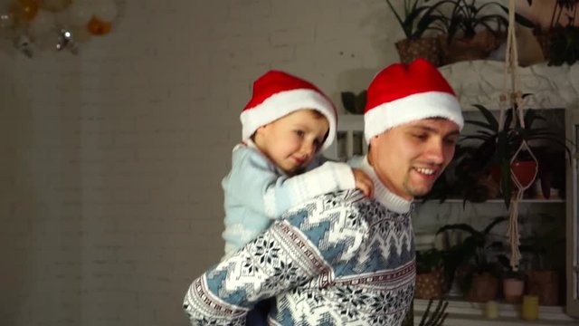 Slow motion piggy back ride of excited loving dad in knitted Christmas sweater and little toddler son in Santa Claus hat