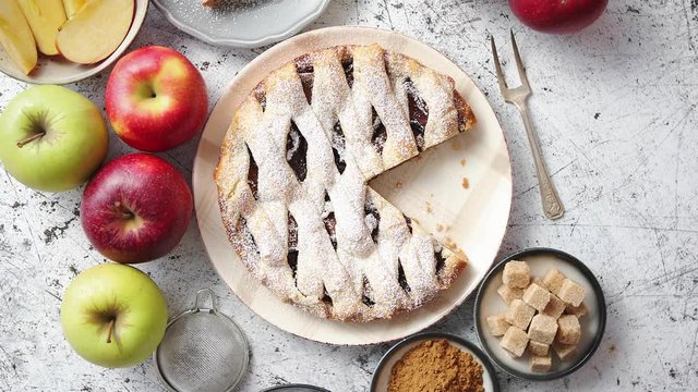 Fresh baked apple pie with cutted slice on small plate. With ingrediends on side. Fresh fruits, brown sugar cubes, cinnamon. Sprinkled with powder sugar.