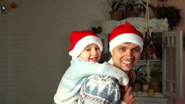 Funny piggy back ride in Santa Claus hats of loving father and cute toddler son in domestic family Christmas atmosphere