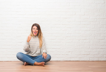 Young adult woman sitting on the floor over white brick wall at home angry and mad raising fist frustrated and furious while shouting with anger. Rage and aggressive concept.