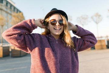 Outdoor portrait of a smiling teen girl 13, 14 years old in a hat and sunglasses. City background, golden hour.