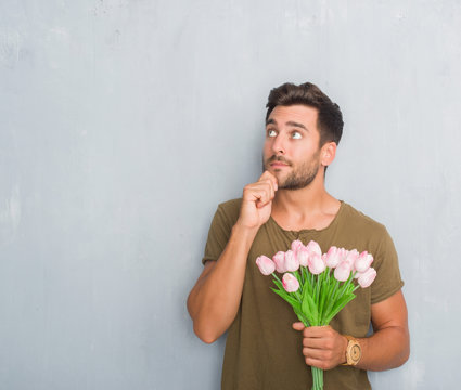 Handsome young man over grey grunge wall holding flowers bouquet serious face thinking about question, very confused idea