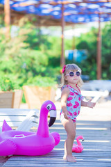 Baby girl on beach with pink flamingo dressed in stylish swimming wear posing on wooden floor. Scene on vacation on sea side of a cute lady smiling and enjoy life time childhood infancy.
